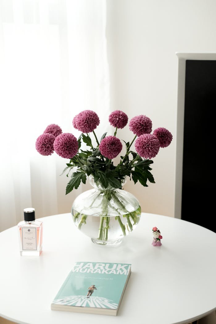 Beautiful pink flowers in a glass vase, accompanied by a book and perfume on a white tabletop.